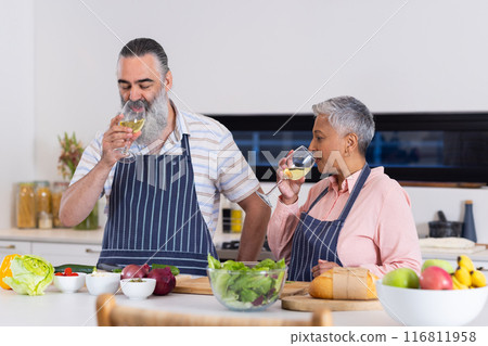 Senior couple in kitchen drinking wine while preparing fresh vegetables and bread 116811958