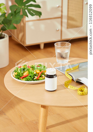 High angle shot at the wooden table top in the room of living, a dish of vegetable salad, measuring tape, glass of water, opened book and a bottle without label placed above. Blank space for text High angle shot at the wooden table top in the room of living, a dish of vegetable salad, measuring tape, glass of water, opened book and a bottle without label placed above. Blank space for text 116812099