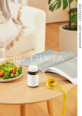 The photograph of living room scene with a wooden table, which displaying some items for losing weight and a blank label supplement bottle. Mock up for advertising with empty space for designing The photograph of living room scene with a wooden table, which displaying some items for losing weight and a blank label supplement bottle. Mock up for advertising with empty space for designing 116812100