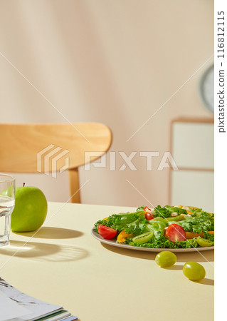 The photo was taken in a room decorated in yellow-beige tones, with a table containing a plate of vegetable salad, an apple and a glass of water. Blank space for showing product and adding text 116812115