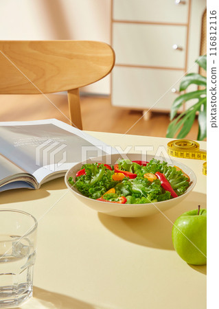 High angle view at a yellow table top, which displaying a dish of vegetable salad, a green apple, a glass of water, measuring tape and an opened book. Blank space on the table for product advertising 116812116
