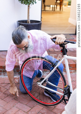 Repairing bicycle, senior man kneeling and fixing wheel outdoors 116812193