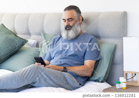 Sitting on bed, man using smartphone and relaxing with medication bottles nearby Sitting on bed, man using smartphone and relaxing with medication bottles nearby 116812281