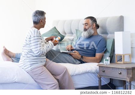 Senior woman giving medication to man using laptop while resting in bed 116812282