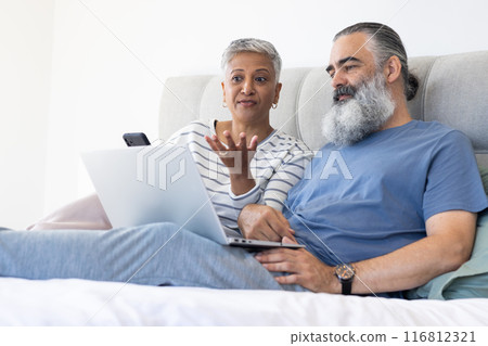 Using laptop and smartphone, senior couple relaxing on bed and discussing something Using laptop and smartphone, senior couple relaxing on bed and discussing something 116812321