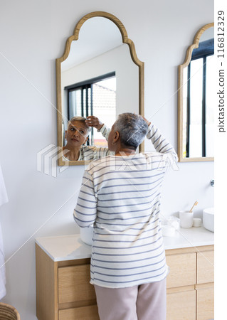 Senior woman looking in mirror and adjusting hair in bathroom Senior woman looking in mirror and adjusting hair in bathroom 116812329