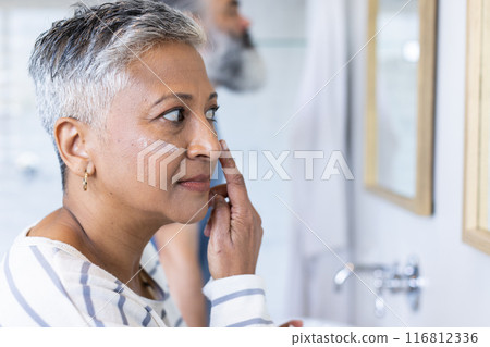 Applying face cream, senior woman focusing on skincare routine in bathroom 116812336