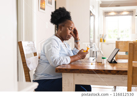 Sad senior african american woman holding pills and using tablet at home Sad senior african american woman holding pills and using tablet at home 116812530