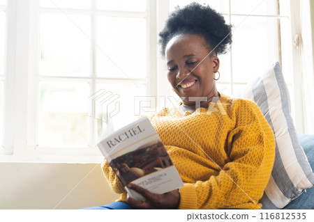 Happy senior african american woman lying on couch and reading book at home 116812535