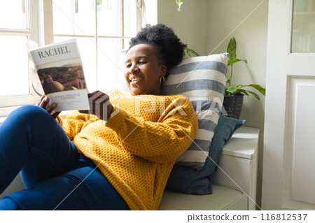 Happy senior african american woman lying on couch and reading book at home 116812537