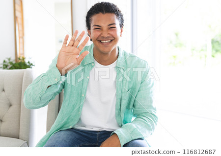 Young biracial man waves cheerfully during a video call, seated indoors with copy space 116812687