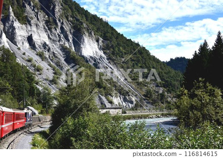 An Albula Line train runs in front of a large exposed monolithic limestone cliff. An Albula Line train runs in front of a large exposed monolithic limestone cliff. 116814615