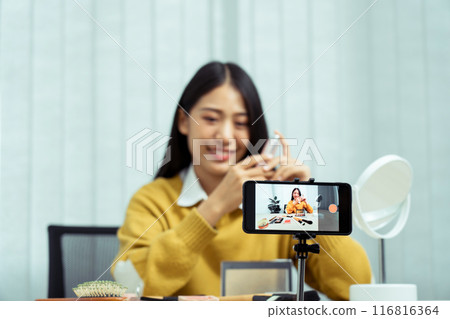 Young asian woman vlogging herself about beauty products with making a video for her blog on cosmetics using phone camera at home. 116816364