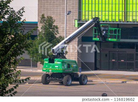 Calgary, Alberta, Canada. Jul 20, 2024. A green telescopic boom lift is positioned in front of a commercial building, with its platform elevated for maintenance work. 116819602