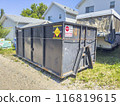 Calgary, Alberta, Canada. Jul 20, 2024. A large black dumpster placed in a residential area, next to white houses and a covered trailer with warning signs on the dumpster. 116819615