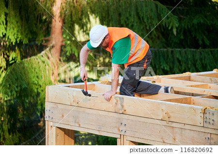 Carpenter constructing two-story wooden frame house near the forest. Man hammering nails into structure while wearing protective helmet and construction vest. Concept of modern ecological construction 116820692