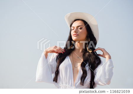 A beautiful woman in a white hat and white dress against a blue sky in summer 116821039