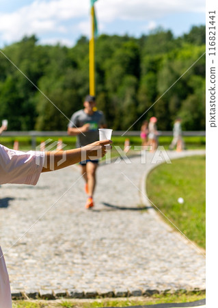 A volunteer in a white dress holds out a plastic cup of water to a triathlon runner. 116821441