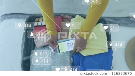 Image of media notifications over biracial woman with smartphone packing suitcase Image of media notifications over biracial woman with smartphone packing suitcase 116823670