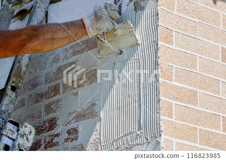 Using notched trowel, construction worker applies an even layer of adhesive to surface of facade. Using notched trowel, construction worker applies an even layer of adhesive to surface of facade. 116823985
