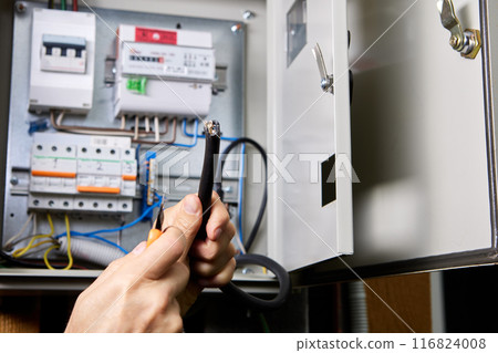 Removing PVC sheath of an electrical cable with retractable utility knife, against background of an electric switchboard. 116824008