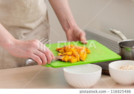 A woman adds chopped peach to her oatmeal. A woman adds chopped peach to her oatmeal. 116825486