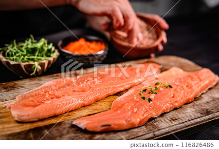 Fresh trout salmon filet with herbs greens on wooden board and chief hands on blurred background. Omega orange fish with rosemary for healthy nutrition Fresh trout salmon filet with herbs greens on wooden board and chief hands on blurred background. Omega orange fish with rosemary for healthy nutrition 116826486
