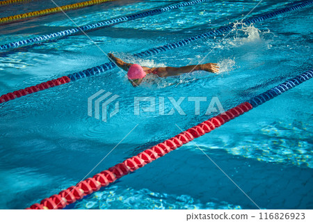 Side view of swimmer glides powerfully through water with butterfly stroke, his focused expression and strong arms making splash in clear blue lane. Side view of swimmer glides powerfully through water with butterfly stroke, his focused expression and strong arms making splash in clear blue lane. 116826923
