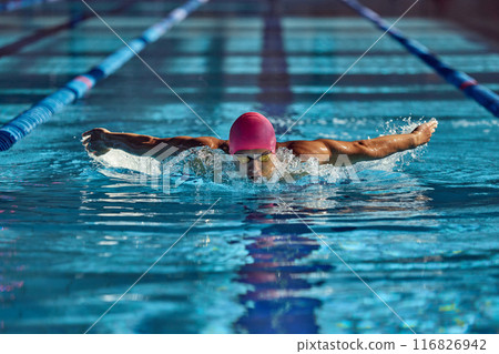 Man, swimmer in pink cap and goggles powers through water with butterfly stroke, his focused expression and strong arms creating dynamic splash in pool. 116826942
