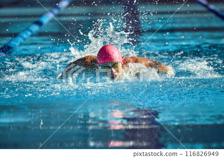 Man, swimmer in pink cap and goggles powers through water with butterfly stroke, his focused expression and strong arms creating dynamic splash in pool. Man, swimmer in pink cap and goggles powers through water with butterfly stroke, his focused expression and strong arms creating dynamic splash in pool. 116826949