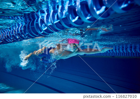 Competitive swimmer, with pink cap and blue trunks, seen underwater during dive, demonstrates his strength and form creating water turbulence as he moves forward. 116827010