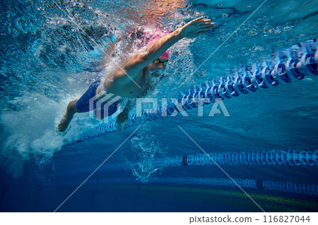 Male athlete swimming, highlighting his focus and form, as he pushes through water in pool, wearing blue swim trunks and goggles. 116827044