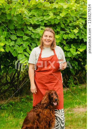 30s Woman in apron with dog tasting pink wine in vineyard. Portrait of pretty young woman holding glass of wine. Happy vintner drinks wine after successful grape harvesting. Harvest time, full body 116827654
