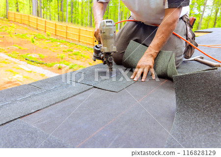 Construction worker by roofer installing new bitumen shingles on roof used air pneumatic nail gun Construction worker by roofer installing new bitumen shingles on roof used air pneumatic nail gun 116828129