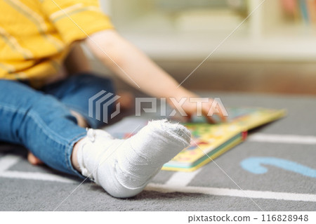 Toddler boy with a bandage or cast on his leg plays with colourful book. Fracture of a foot and finger in children. Human healthcare and medicine concept. focus on the cast Toddler boy with a bandage or cast on his leg plays with colourful book. Fracture of a foot and finger in children. Human healthcare and medicine concept. focus on the cast 116828948