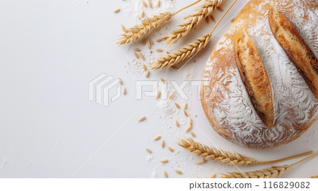 A freshly baked wheat bread loaf with a golden-brown crust is displayed on a white background, surrounded by wheat stalks and scattered flour. 116829082