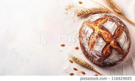 A freshly baked bread loaf made with wheat flour sits on a white background with wheat stalks and almonds scattered around it. 116829085