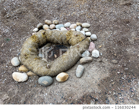 This image depicts an outdoor drainage system protected by erosion control measures. The drain is surrounded by a circular barrier made of natural fiber rolls and large river rocks. The setup ensures 116830244