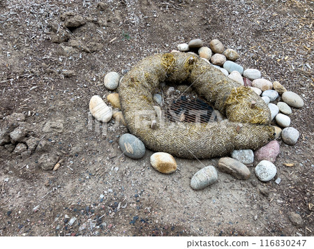 This image depicts an outdoor drainage system protected by erosion control measures. The drain is surrounded by a circular barrier made of natural fiber rolls and large river rocks. The setup ensures 116830247