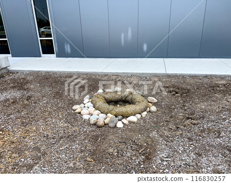 This image depicts an outdoor drainage system protected by erosion control measures. The drain is surrounded by a circular barrier made of natural fiber rolls and large river rocks. The setup ensures 116830257