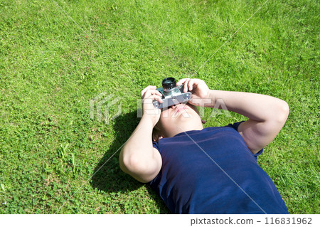 Boy is lies on green grass and takes shoot by old and vintage photo camera. Outdoors. Copy space. 116831962