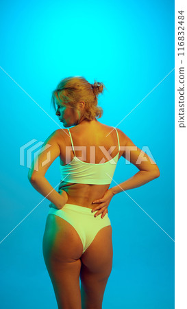 Rear view of blonde woman in white underwear posing holding hands on hips in neon light against gradient blue background. 116832484