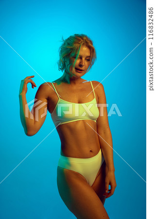 Portrait of middle aged beautiful woman posing in lingerie shows her fit, smooth body in neon light against gradient blue background. Portrait of middle aged beautiful woman posing in lingerie shows her fit, smooth body in neon light against gradient blue background. 116832486