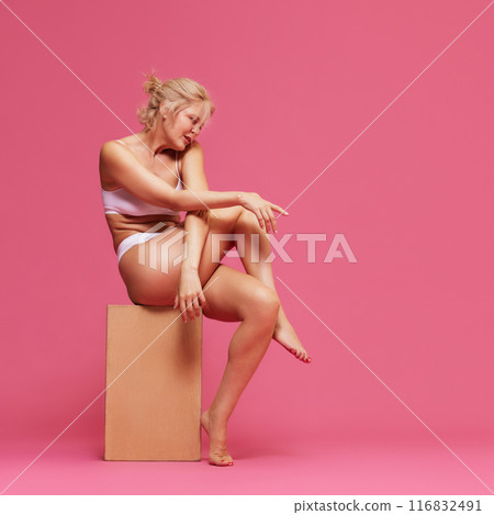 Poised woman, in simple white lingerie, sitting on wooden block crossed hands and legs against pink studio background. Poised woman, in simple white lingerie, sitting on wooden block crossed hands and legs against pink studio background. 116832491