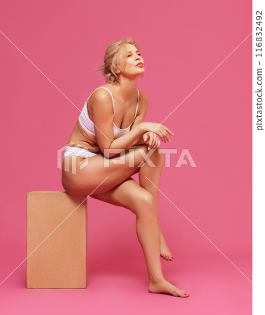 Thoughtful woman in white lingerie sits gracefully on wooden block, exuding confidence and elegance against pink background. 116832492