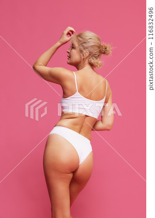 Rear view of blonde woman with fit, toned body in white lingerie posing raising hand against pink background. 116832496
