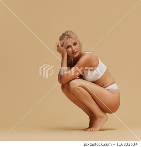 Blonde woman in white lingerie crouches gracefully and looking at camera with confidence against pastel studio background. 116832534