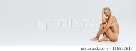 Banner. Posed in nude lingerie, blonde woman sits with one hand on her head against white studio background with negative space. Banner. Posed in nude lingerie, blonde woman sits with one hand on her head against white studio background with negative space. 116832613