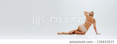 Banner. Rear view of woman in beige lingerie lying, posing on floor against white studio background, exuding beauty an femininity. Banner. Rear view of woman in beige lingerie lying, posing on floor against white studio background, exuding beauty an femininity. 116832615