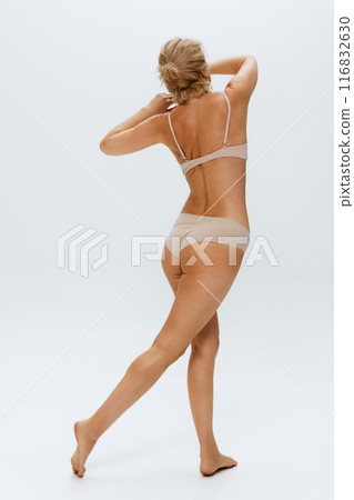 Back view of blonde woman wearing beige inner wear stretching her arms, legs and spine against white studio background. 116832630